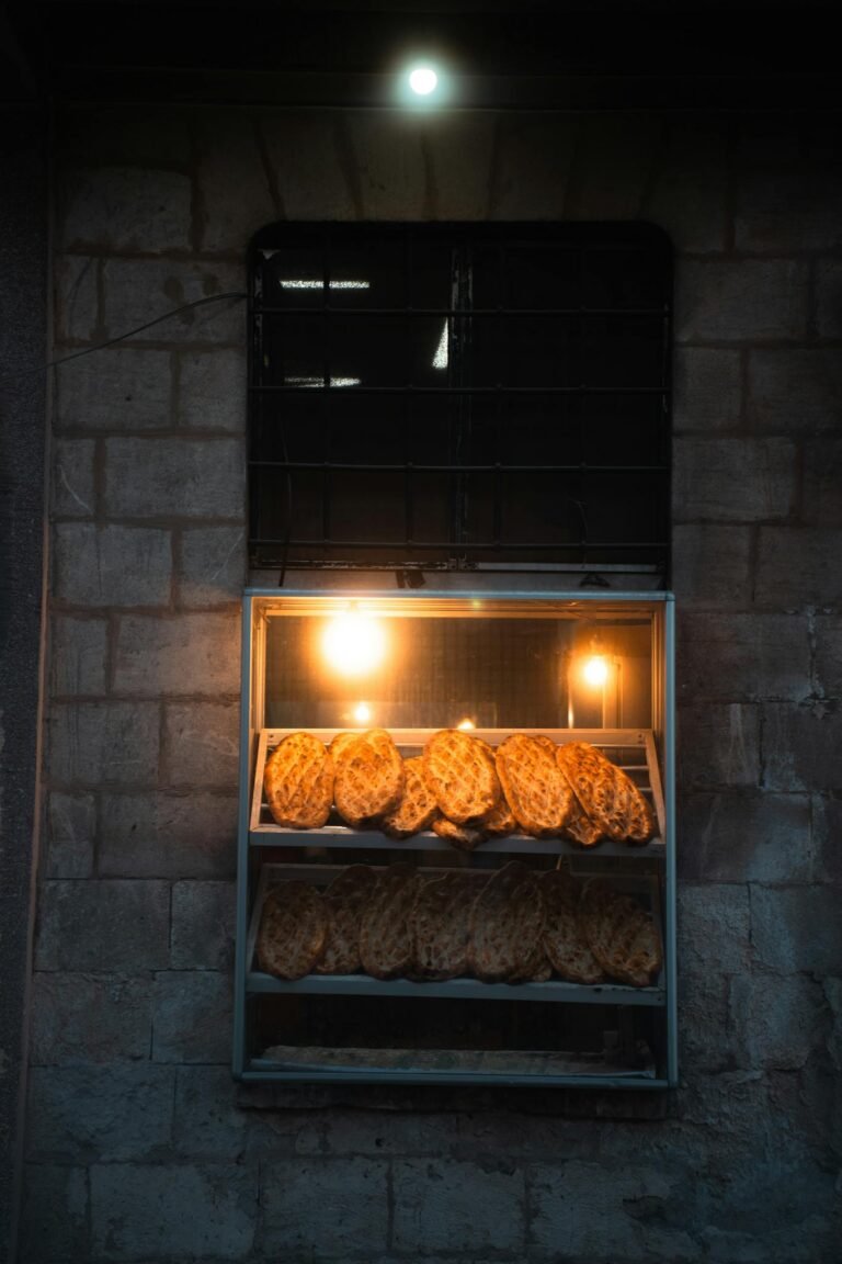Loaves of bread showcased in a bakery window, warmly lit against brick walls at night.